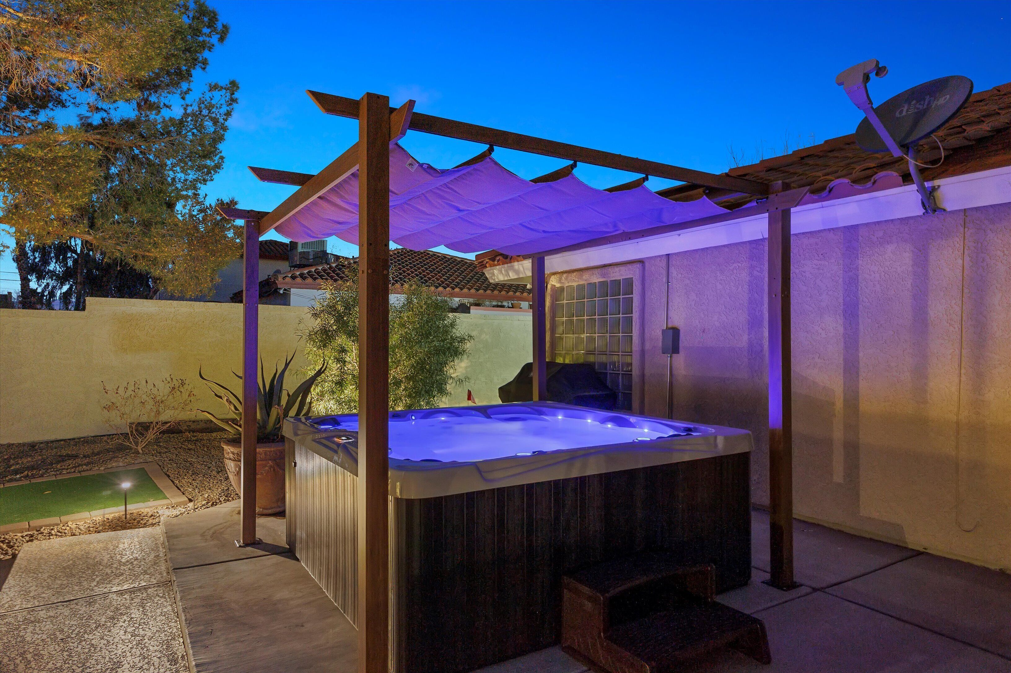 A hot tub under a pergola with a satellite dish, surrounded by a garden and a house.
