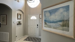 A hallway with a white door, a framed painting of a beach scene, a decorative light fixture, and a small shelf with a plant. - Sun Palm Savannah at Windsor Palms Resort (Kissimmee)
