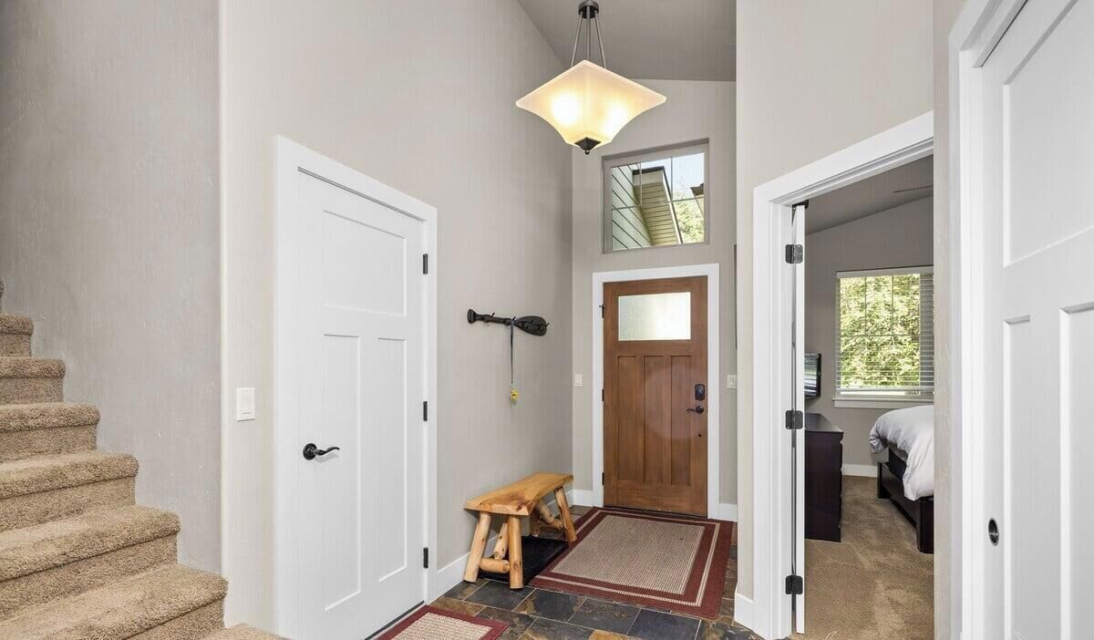 A hallway with a staircase, a wooden bench, a door with a mirror, and a ceiling light fixture.