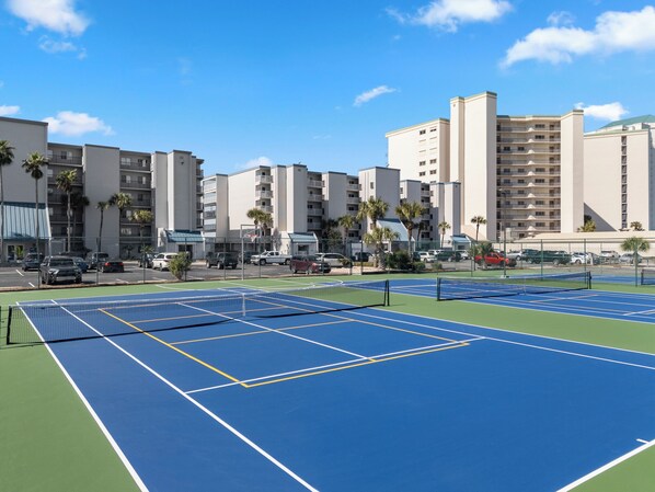 Una cancha de tenis con red, rodeada por una cerca, y contigua a un estacionamiento con coches.