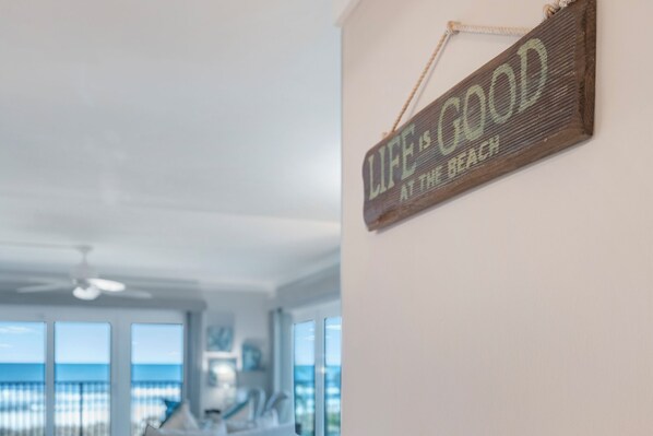 Una habitación con un ventanal grande con vistas a la playa, un ventilador de techo y un letrero de madera en la pared que dice 'LIFE IS GOOD AT THE BEACH'.