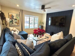 Una sala de estar moderna con un sofá de color oscuro, una mesa de centro de cristal y un televisor de pantalla plana grande montado en la pared.