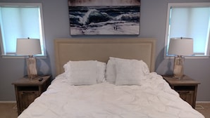 A bedroom with a large bed, two bedside tables with lamps, a headboard, and a painting of a seascape on the wall.