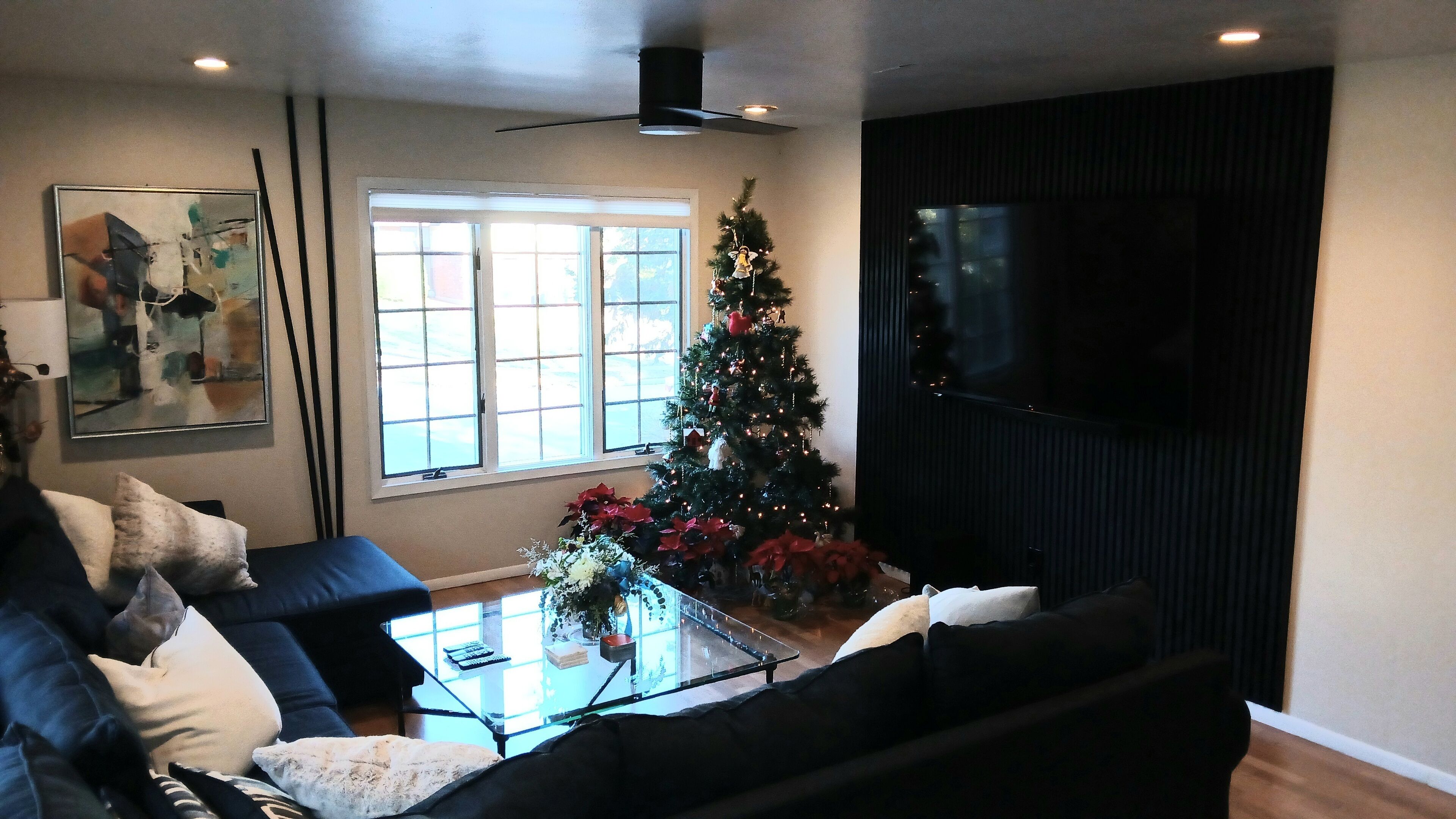 A living room with a black couch, a glass coffee table, a Christmas tree, and a large flat-screen TV.
