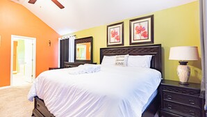 Une chambre avec un grand lit, une table de chevet avec une lampe, un ventilateur de plafond et deux tableaux floraux encadrés accrochés au mur.