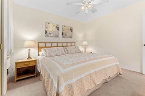 A bedroom with a bed, two bedside lamps, a ceiling fan, and two wall-mounted artworks. - Koa Lagoon 506 by Maui Rental Group (Kihei)