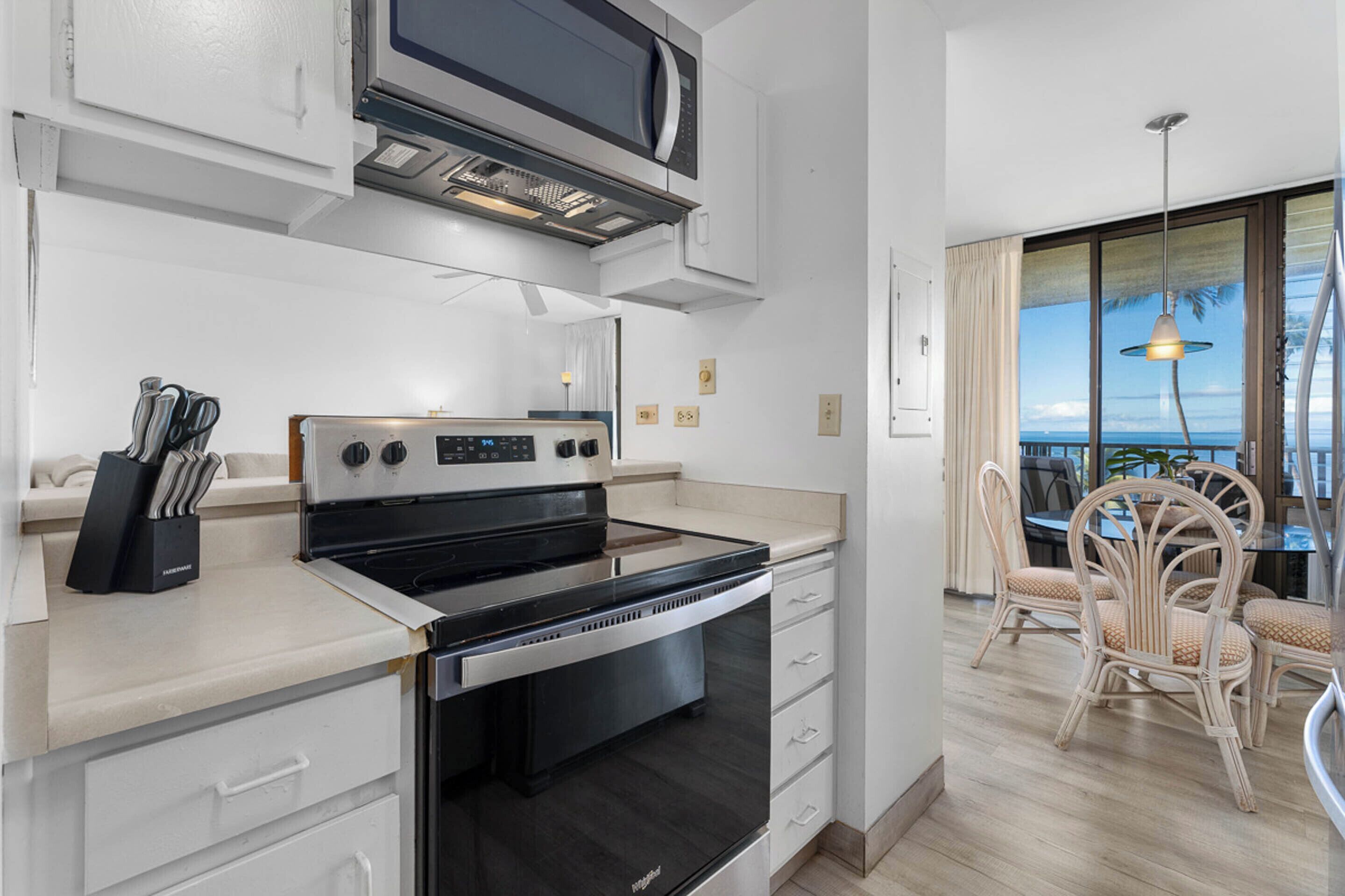 A modern kitchen with a built-in oven and microwave, a dining area with wicker chairs, and a large window offering a view of the ocean.