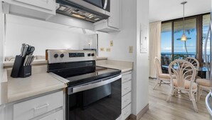A modern kitchen with a built-in oven and microwave, a dining area with wicker chairs, and a large window offering a view of the ocean. - Koa Lagoon 506 by Maui Rental Group (Kihei)