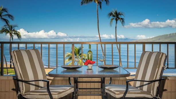 A balcony with two chairs, a table, and a bottle of wine. - Koa Lagoon 506 by Maui Rental Group (Kihei)