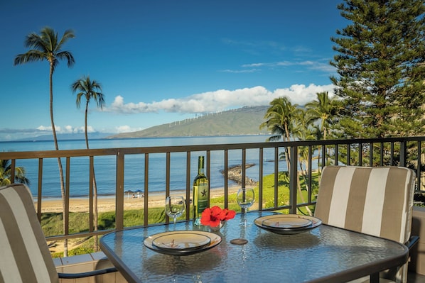 A table with a glass top, two chairs, a bottle, and a glass of wine on a balcony overlooking a beach with palm trees and a view of a distant island. - Koa Lagoon 506 by Maui Rental Group (Kihei)