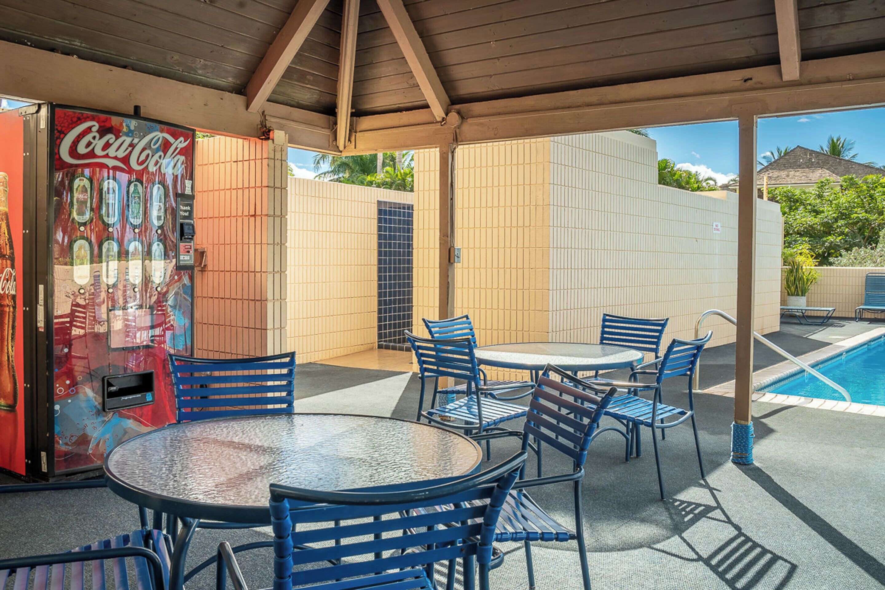Une terrasse extérieure avec une piscine, des chaises en métal bleues et un distributeur automatique de Coca-Cola.