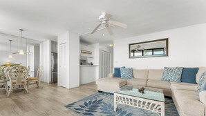 A modern living room with a beige sofa, a glass coffee table, and a dining area with wicker chairs. - Koa Lagoon 506 by Maui Rental Group (Kihei)
