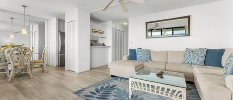 A modern living room with a beige sofa, a glass coffee table, and a dining area with wicker chairs.