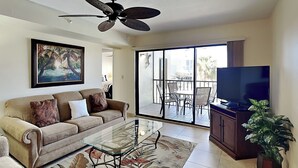 A living room with a sofa, a glass coffee table, a ceiling fan, a TV, and a sliding glass door leading to an outdoor area with chairs and a table. - Santa Maria 208 by Distinctive Beach Rentals (Fort Myers Beach)