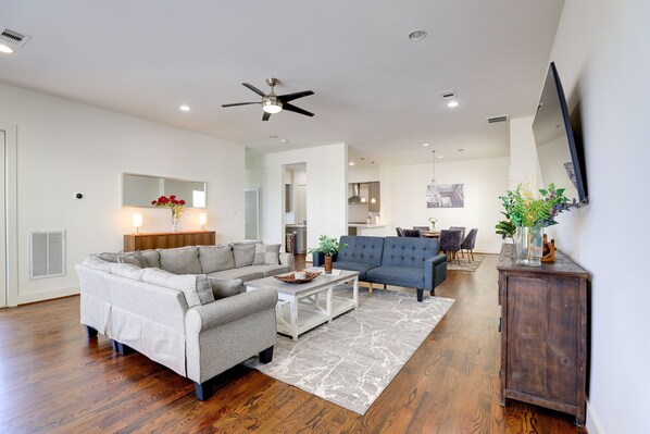 A spacious living room with a sectional sofa, a blue armchair, and a coffee table. There is a wooden cabinet and a flat-screen TV mounted on the wall. - 3-Story Home Near Museums, Nightlife & Downtown! (Houston)