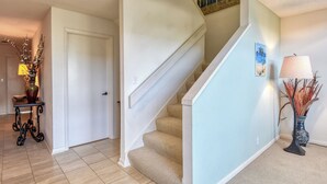 Una escalera con peldaños cubiertos de alfombra color beige y paredes blancas. Hay una puerta a la izquierda y una lámpara a la derecha.