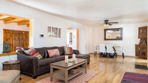 A living room with a brown leather sofa, a wooden coffee table, a dining table with chairs, and a ceiling fan. - Rio Grande Retreat by Kokopelli Property Management (Santa Fe)