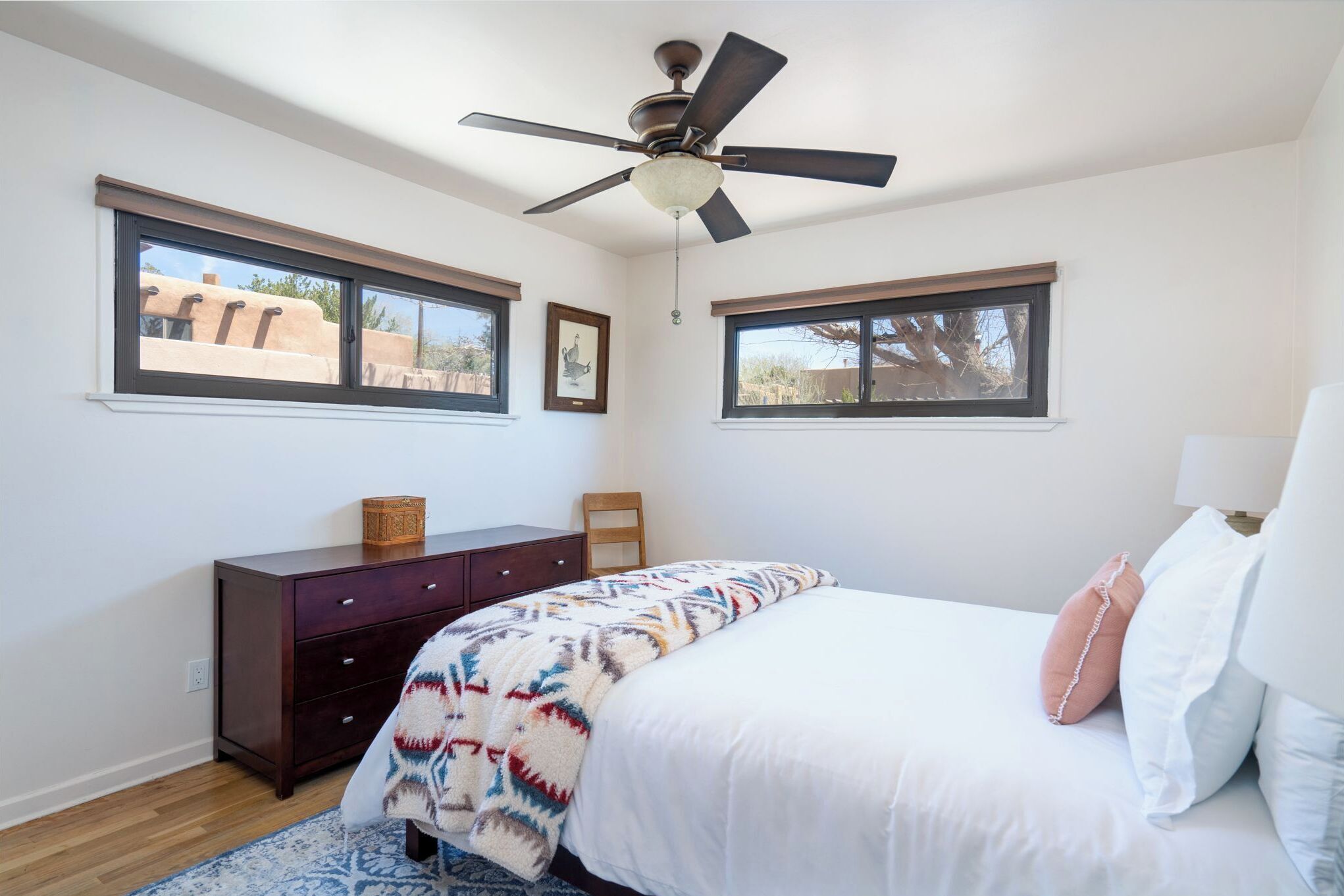 Un dormitorio con cama, cómoda, ventilador de techo y dos ventanas.