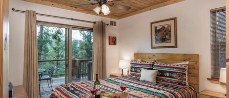 A bedroom with a wooden ceiling, a bed with patterned bedding, a nightstand, a window with blinds, and a balcony with a chair.