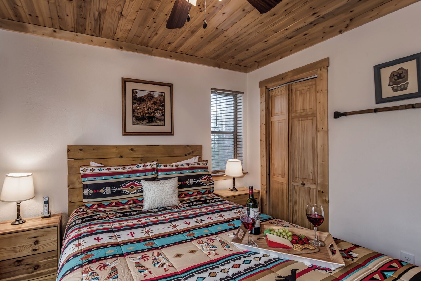 A bedroom with a wooden headboard, a bed with patterned bedding, a bedside table with a lamp, a window with blinds, and a wooden door.