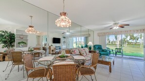 A spacious living room with a dining area, large windows, and a view of a golf course. - Vista Verde North 2-110 by Distinctive Beach Rentals (St. Petersburg)