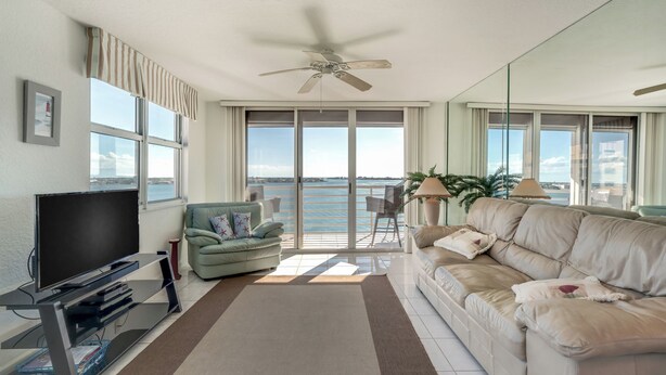 A modern living room with a flat-screen TV, a ceiling fan, a sofa, a chair, and a view of the sea through large windows. - Bahia Vista 17-881 by Distinctive Beach Rentals (St. Petersburg)