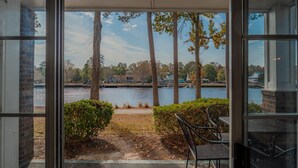 Vista desde una habitación con puerta de vidrio, con vistas a un lago y casas y árboles al fondo.