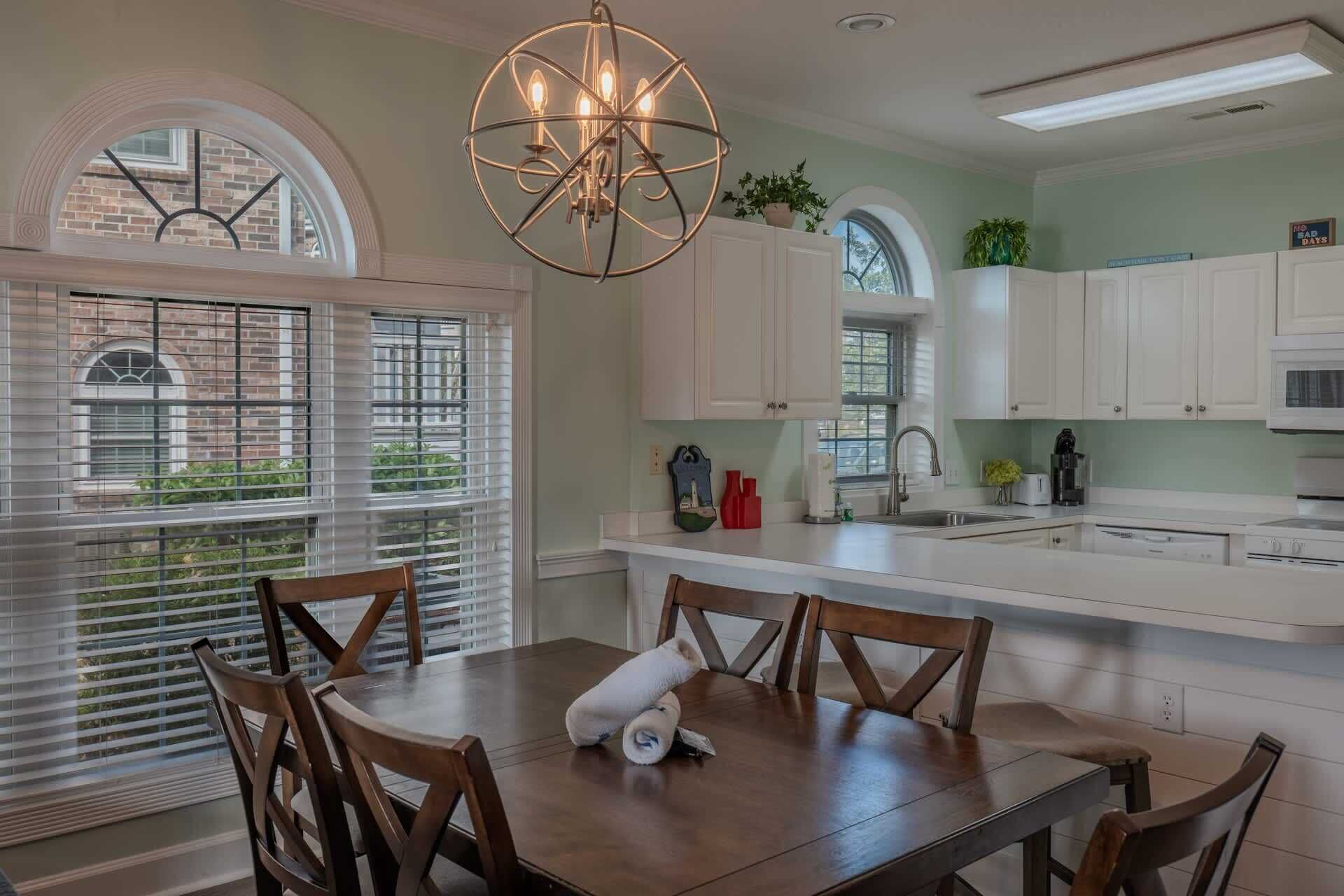 A kitchen with white cabinets, a dining table with chairs, a chandelier, and a view of the outside through the window.
