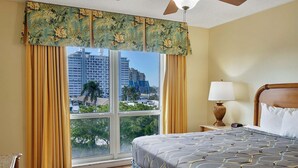 Quarto de hotel com uma grande janela, cama com colcha estampada, mesa de cabeceira com abajur e vista para a cidade e palmeiras.