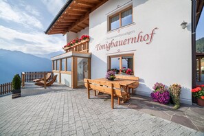 En bygning med et skilt, der viser 'Ferienswohnungen Tonigbauernhof', og en træterrasse med bænke og blomsterkrukker.