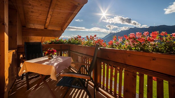 A wooden deck with a table and chairs, a wooden railing, and a view of mountains and flowers.