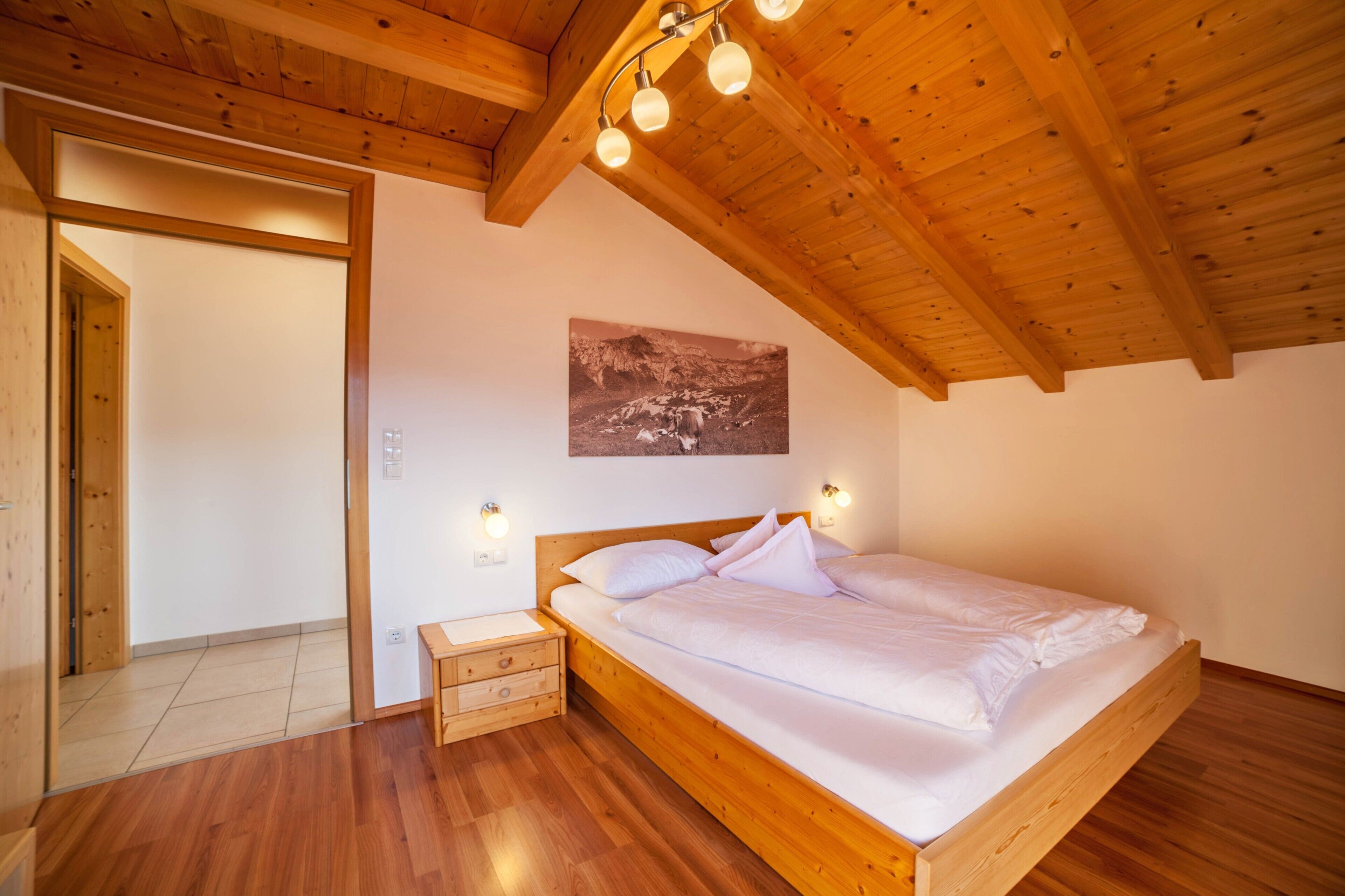 Quarto com cama de madeira, mesa de cabeceira, luminária na parede e quadro emoldurado.