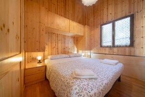 Habitación con paneles de madera, cama, mesita de noche, lámpara y ventana con cortinas estampadas.