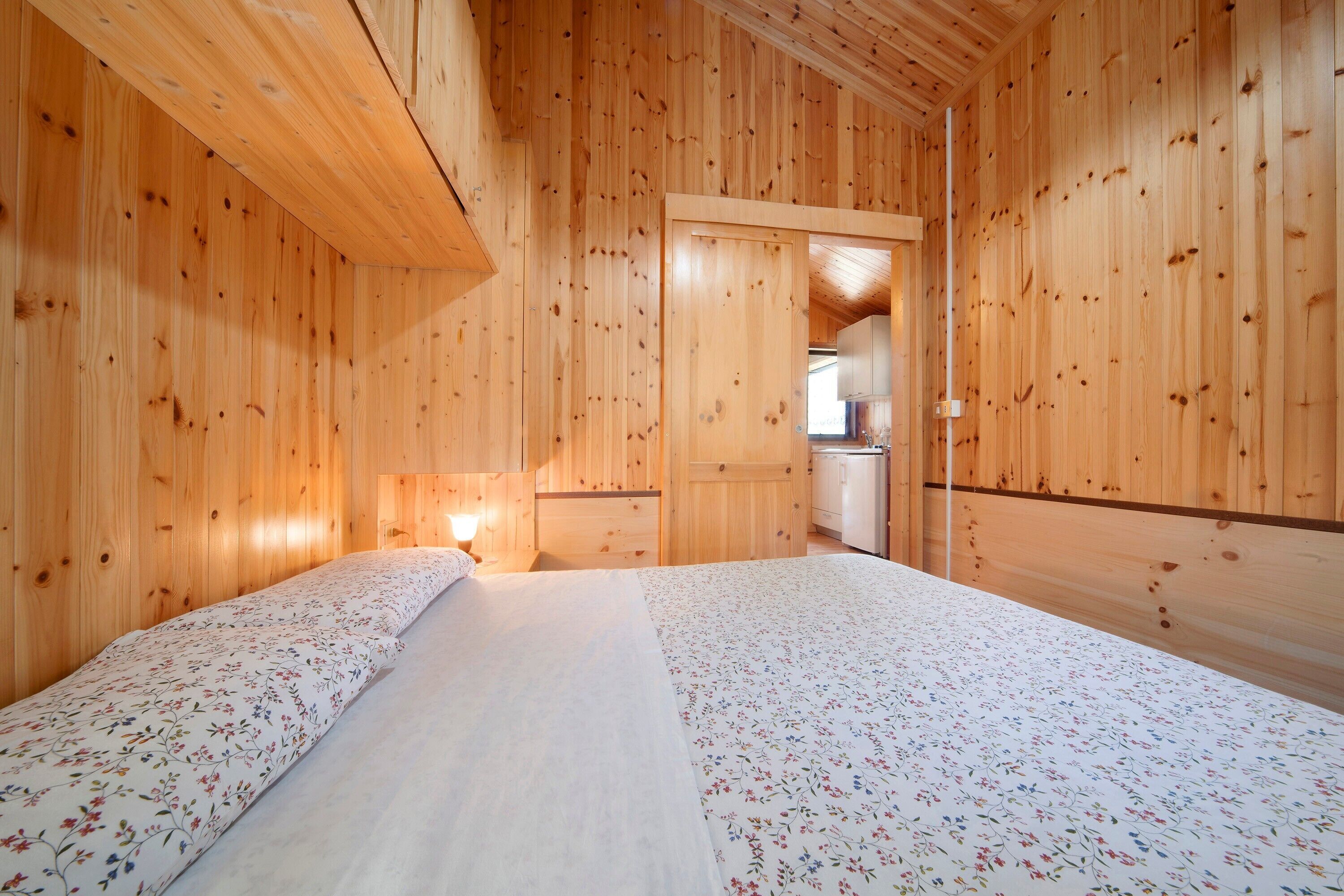 A wooden cabin room with a bed, a small kitchenette, and a wooden ceiling.
