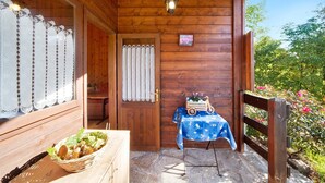 Una cabaña de madera con puerta de madera, una cesta de mimbre con alimentos, una mesa con mantel azul y un banco de madera.