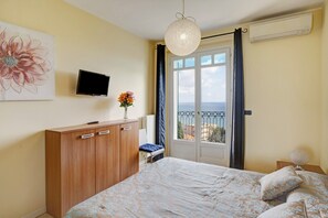 A bedroom with a bed, a TV, a window with curtains, a radiator, and a wall-mounted artwork.
