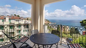 Ein Balkon mit rundem Tisch und Stühlen, der Blick auf die Küste mit Gebäuden und einem klaren Himmel bietet.