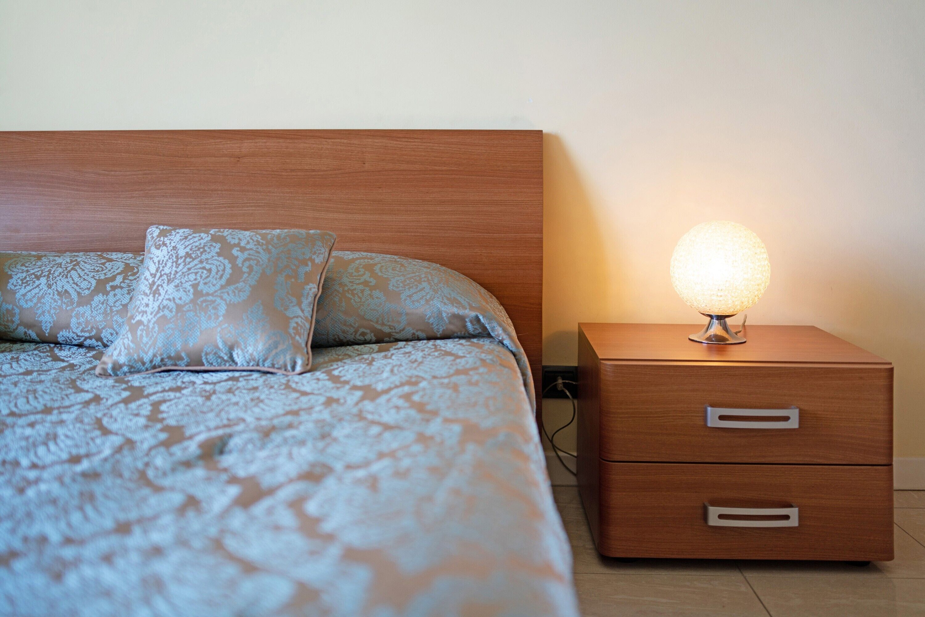 Een bed met behangpatroon, een nachtkastje met lades en een wandlamp.