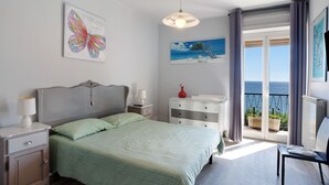 A bedroom with a bed, a nightstand, a dresser, a chair, a window with a view of the sea, and artwork on the walls. - Apartment "Raineri" with Sea View, Wi-Fi, A/C and Balcony (IMPERIA)