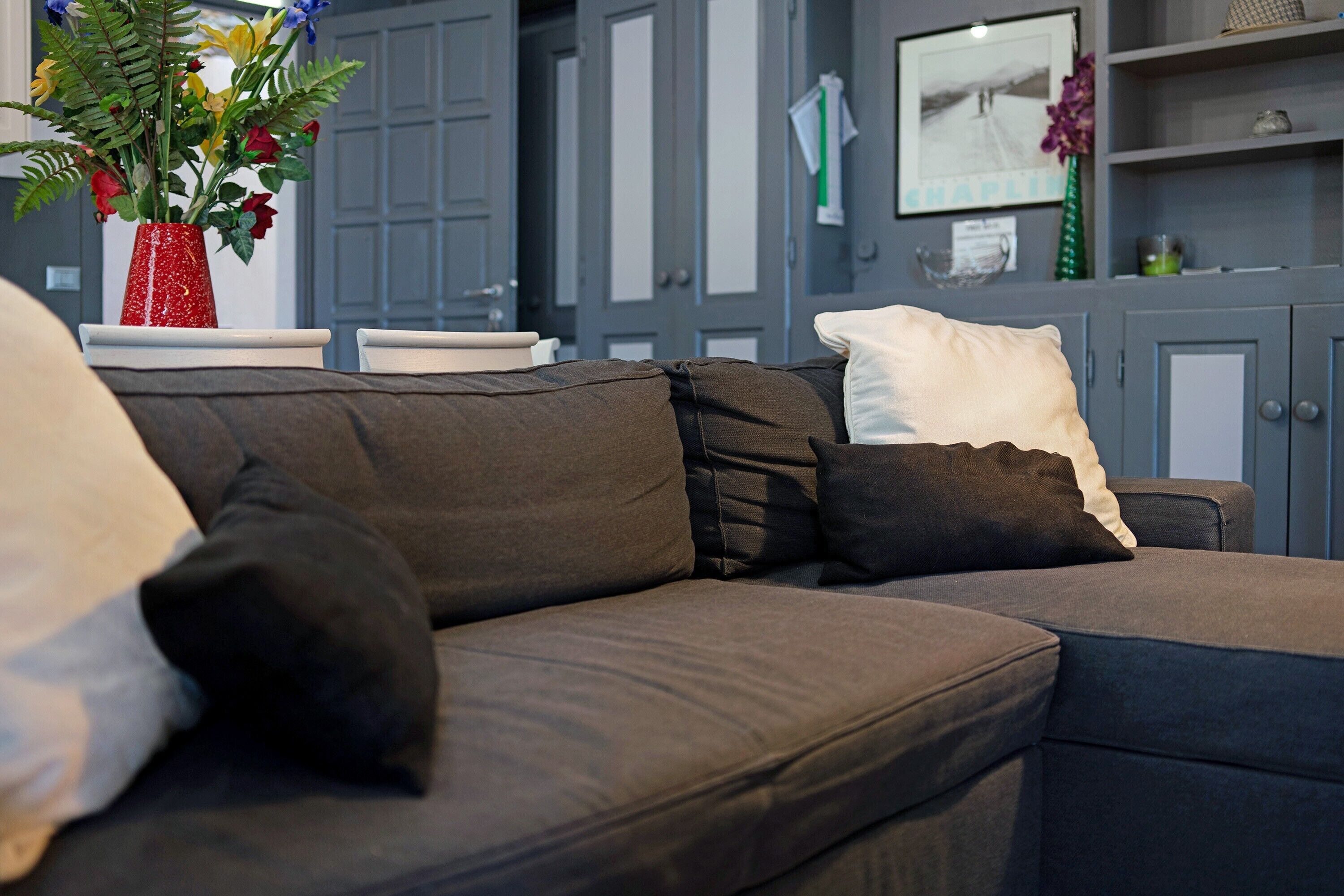 A modern living room with a dark grey sofa, a red vase with flowers, and a framed picture on the wall.
