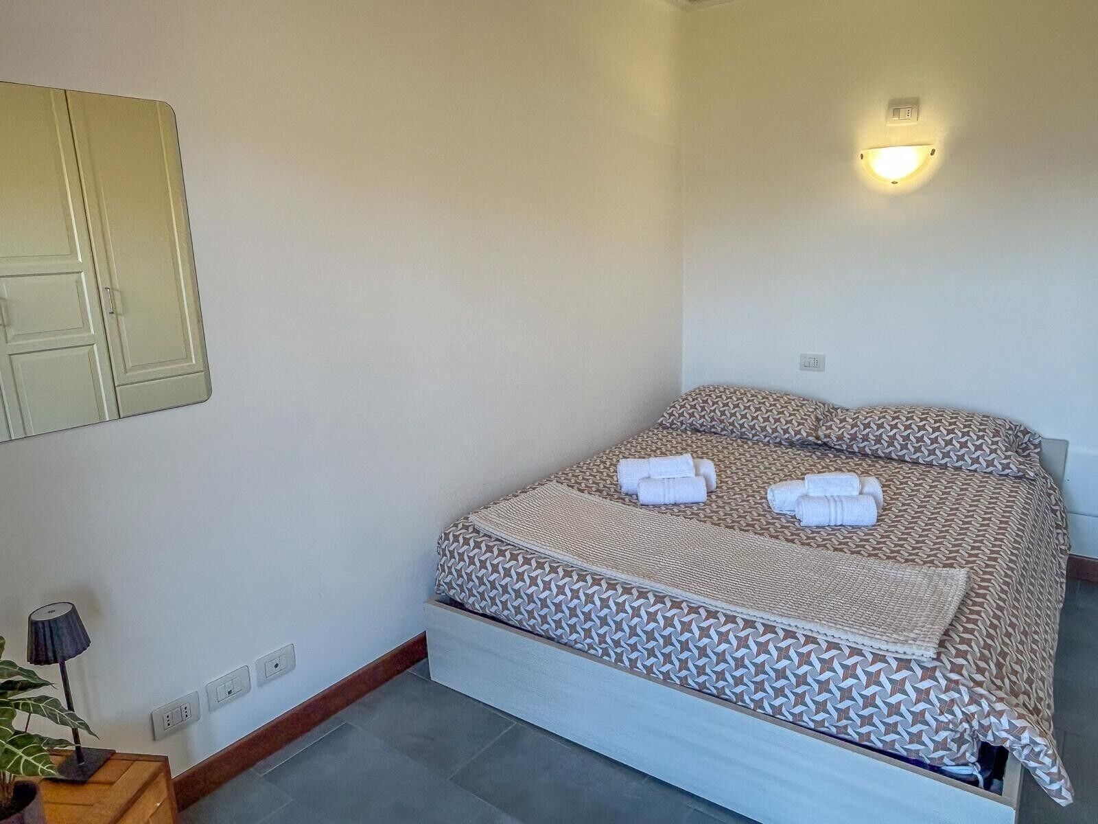 Una cama individual con una colcha estampada, dos toallas ordenadamente dobladas, una lámpara de noche y un aplique de pared.