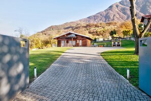 A wooden building with a paved pathway leading to it, surrounded by greenery and mountains.