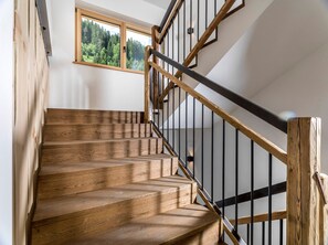 Una escalera de madera con barandilla de metal negro, una ventana con vista al bosque y una pared blanca.