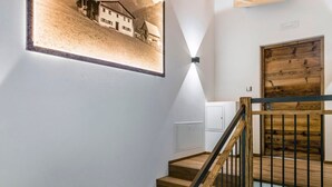 Un escalier avec des marches en bois et des rampes noires, un luminaire fixé au mur et un tableau représentant un bâtiment.