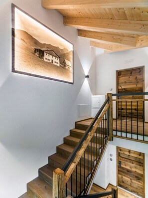 Una escalera con escalones de madera y barandillas negras, una lámpara fijada a la pared y un cuadro con la imagen de un edificio.