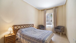 Una camera da letto con un letto, un comodino con lampada, due sedie e una finestra con tende.