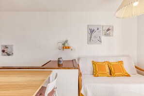 Una habitación con una mesa de madera, un sofá blanco con cojines amarillos, una pequeña estantería con una planta y dos ilustraciones botánicas enmarcadas en la pared.