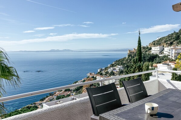 A balcony with a table, chairs, and a cup, overlooking a coastal view with buildings and trees.