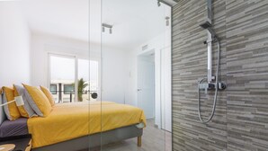 Un dormitorio moderno con separador de vidrio, cama con ropa de cama amarilla, ducha con cabina de vidrio y vistas al exterior.