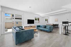 A modern living room with two sofas, a TV, a dining area with chairs, and a kitchen visible through a sliding door.